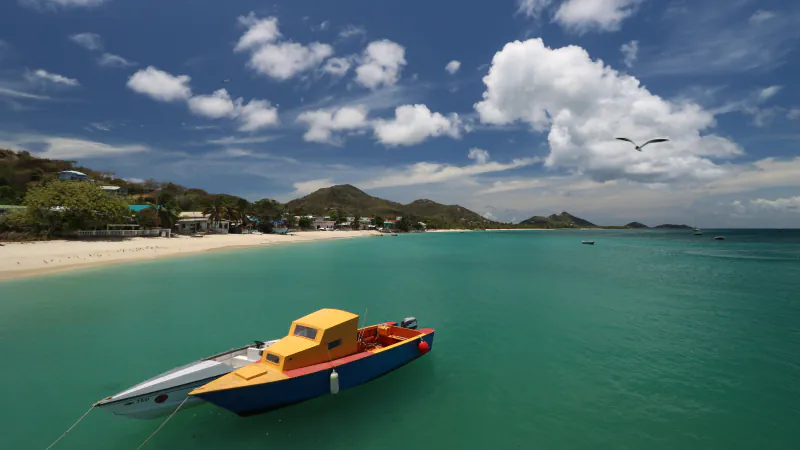 A yellow-cabin boat with a red deck and dark blue hull floats beside a smaller white craft marked TAXI; sandy shore, houses, green hills and clouds fill the background