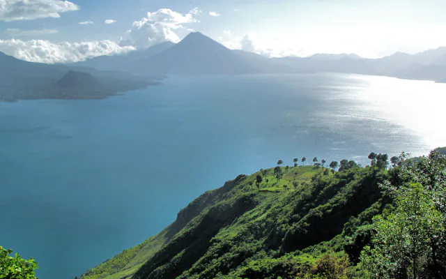 A wide blue lake between green and hazy blue mountains under scattered cloud, with a pointed volcanic peak across the water and a steep green hillside with a few trees in the foreground