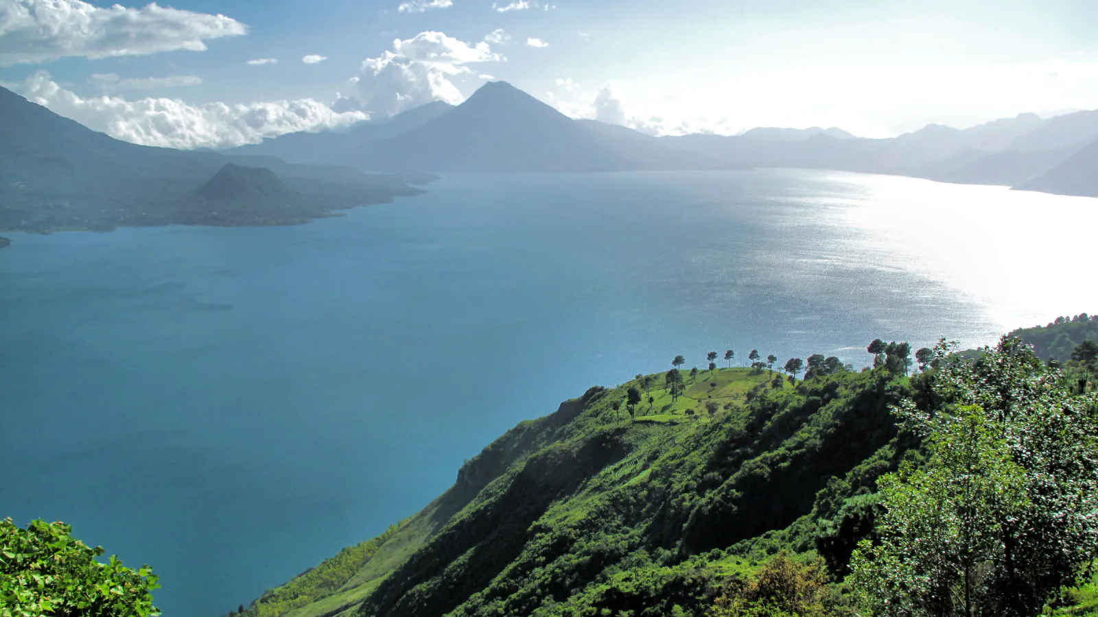 A wide blue lake between green and hazy blue mountains under scattered cloud, with a pointed volcanic peak across the water and a steep green hillside with a few trees in the foreground