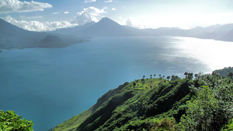 A wide blue lake between green and hazy blue mountains under scattered cloud, with a pointed volcanic peak across the water and a steep green hillside with a few trees in the foreground