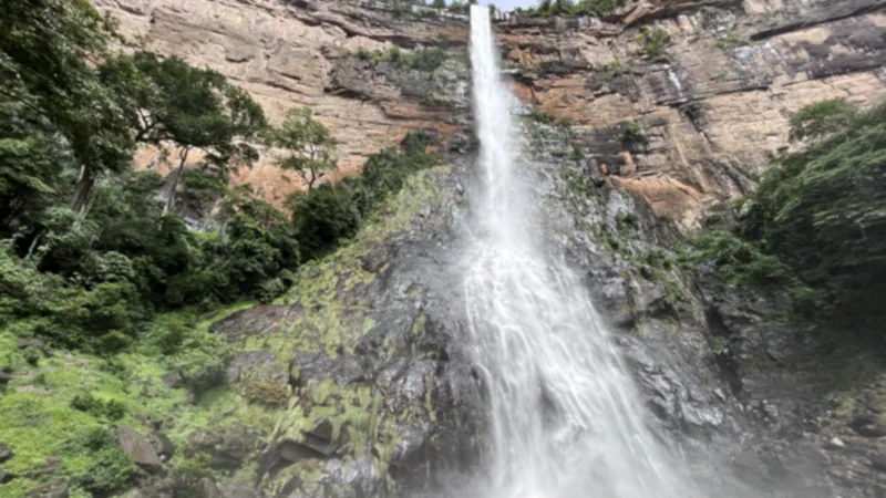A tall, narrow waterfall dropping from the top of a reddish rock cliff, with green forest on the left and dark wet rock below the fall