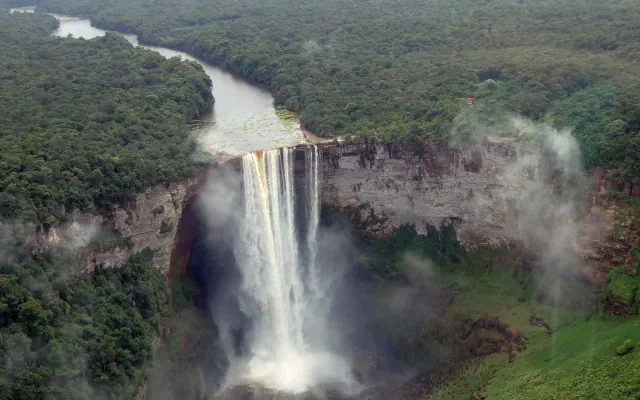 Aerial view of Kaieteur Falls as a broad river drops over a layered cliff into a forested gorge, with mist rising from the base