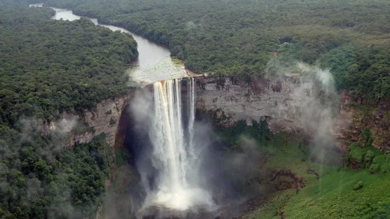 Aerial view of Kaieteur Falls as a broad river drops over a layered cliff into a forested gorge, with mist rising from the base