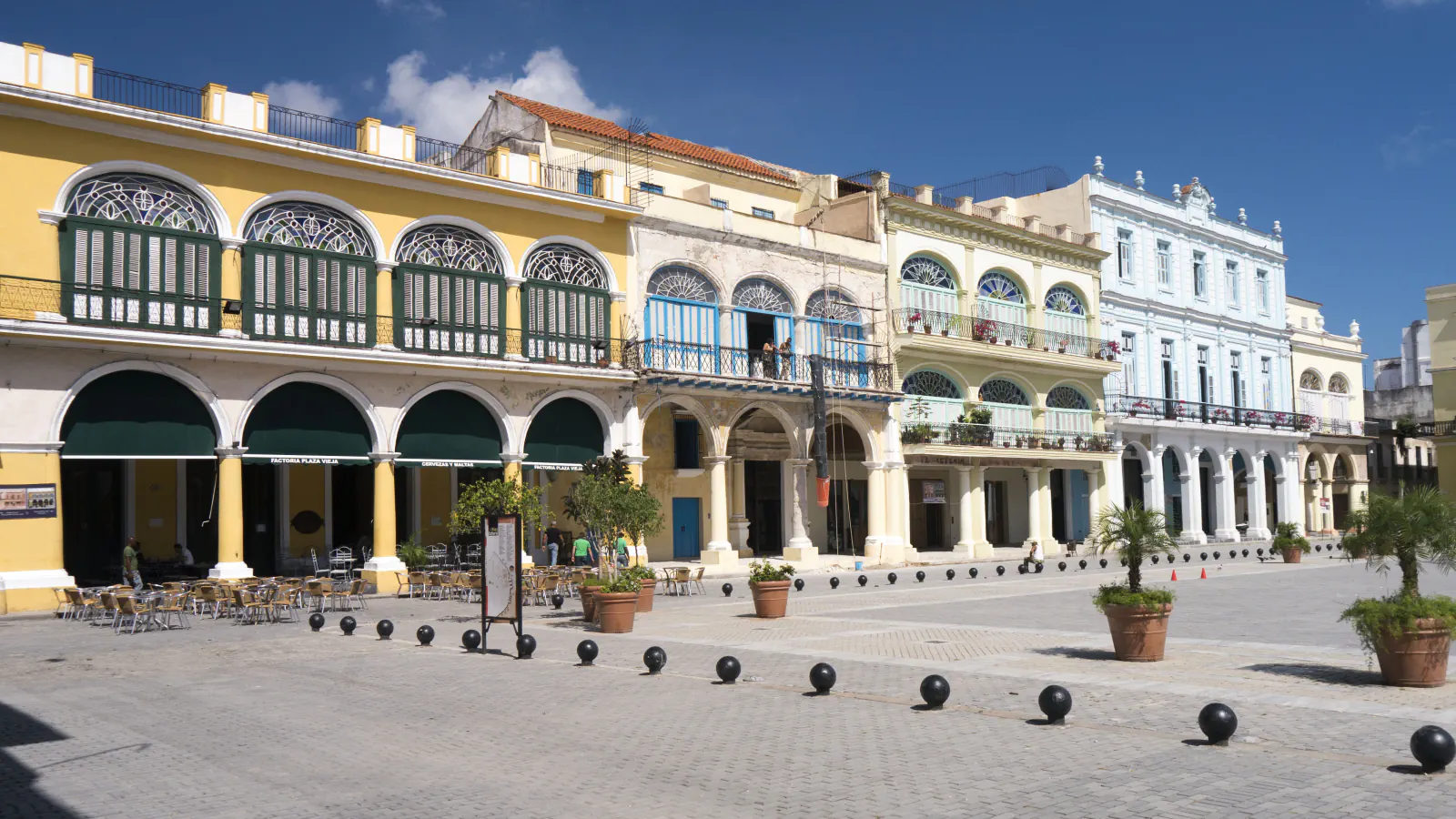 Arcaded houses along one side of Plaza Vieja in Havana: a yellow house with green shutters, a weathered house with blue balconies and pastel façades, on a cobbled square with black stone bollards