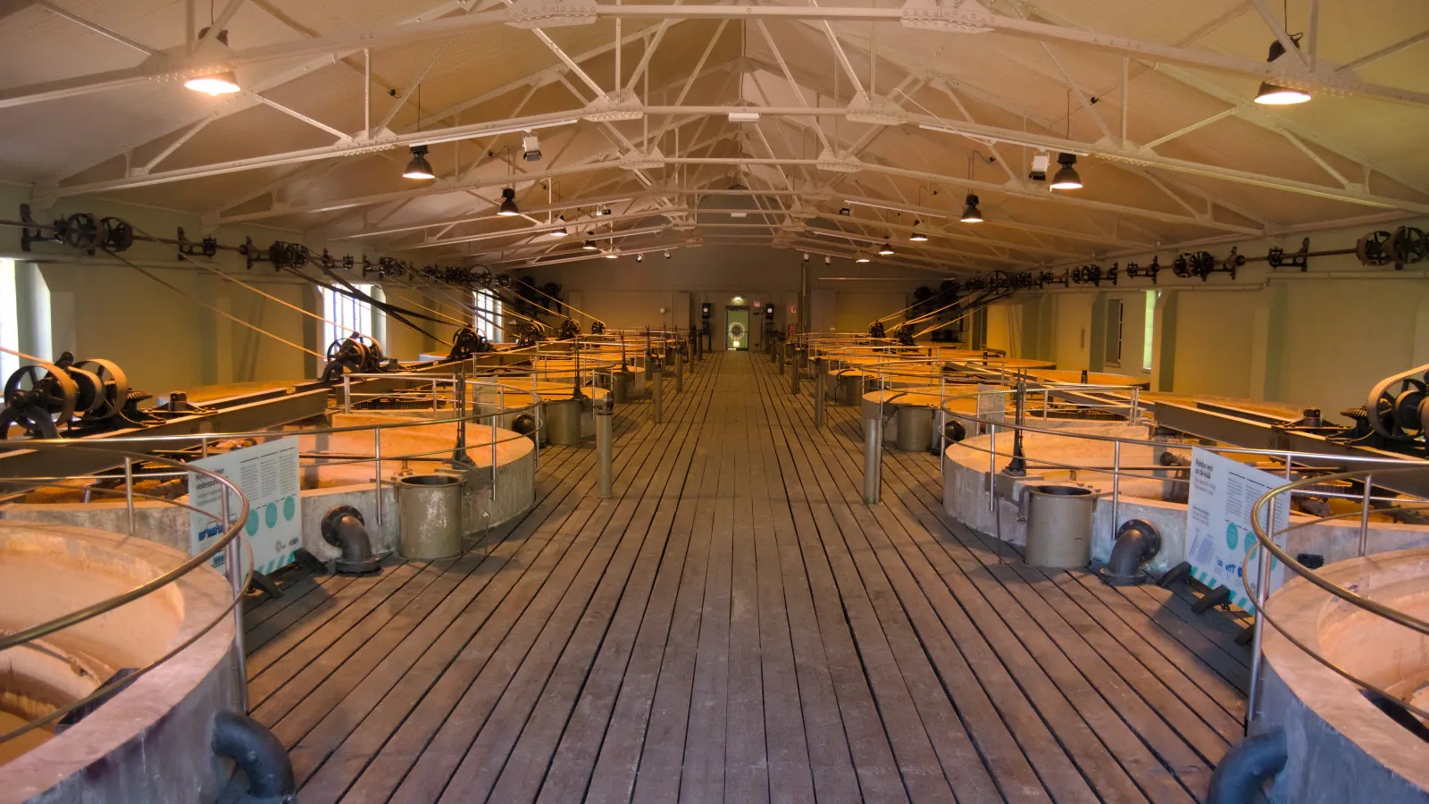 Long hall with a row of round filter basins on each side of a wooden plank walkway, belt drives along the walls and white roof trusses overhead