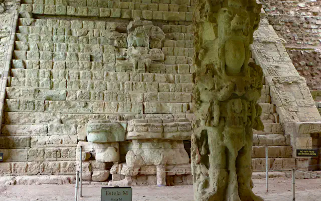 A tall flight of weathered stone steps with rows of carved glyph blocks, a large carved stone stela standing in front on the right and a rope barrier along the base