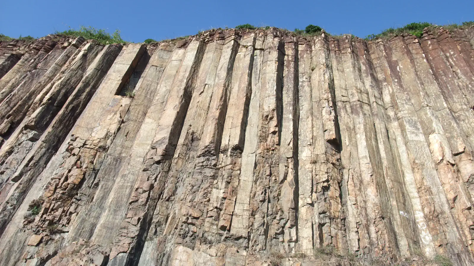 Tall cliff of closely packed, steep rock columns at the East Dam of High Island Reservoir in Hong Kong, with a blue sky above and small green plants along the top