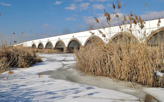 White stone bridge with brown-edged arches seen across a frozen river and tall winter reeds under a blue sky