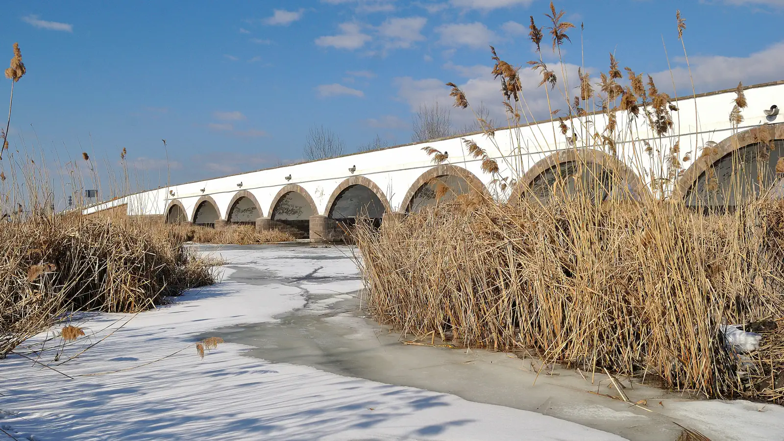White stone bridge with brown-edged arches seen across a frozen river and tall winter reeds under a blue sky