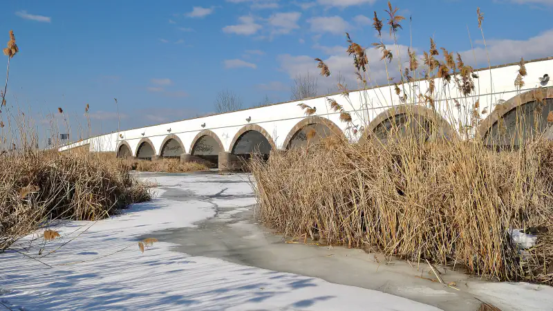 White stone bridge with brown-edged arches seen across a frozen river and tall winter reeds under a blue sky