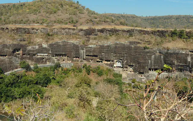 A long dark rock cliff with rows of cave openings above a wooded valley at Ajanta, with scrubby hills rising behind