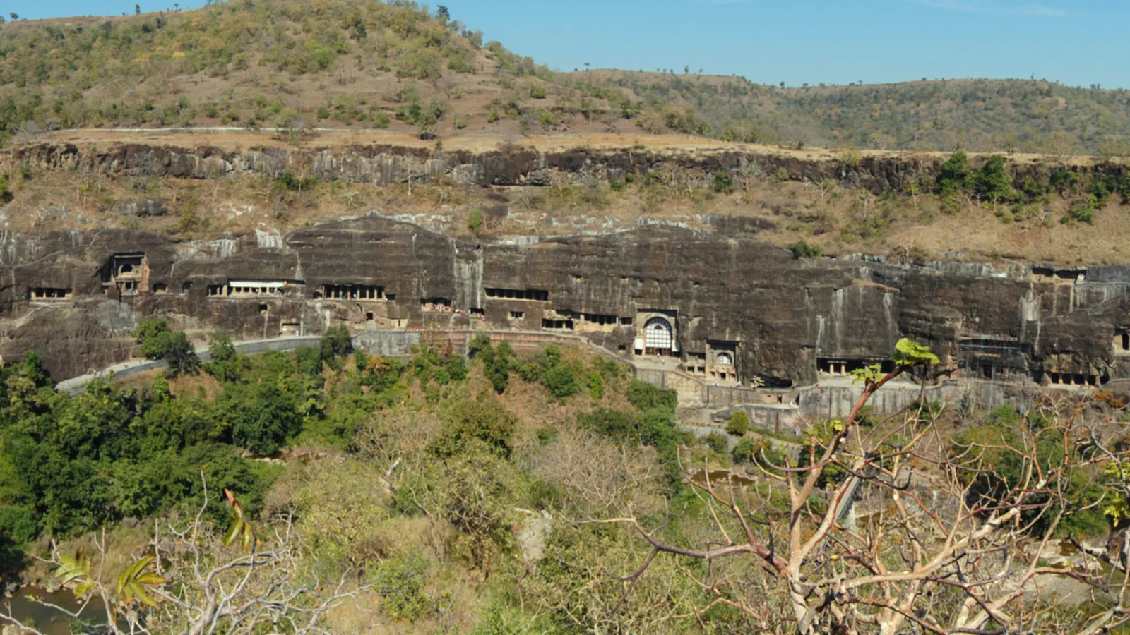 A long dark rock cliff with rows of cave openings above a wooded valley at Ajanta, with scrubby hills rising behind