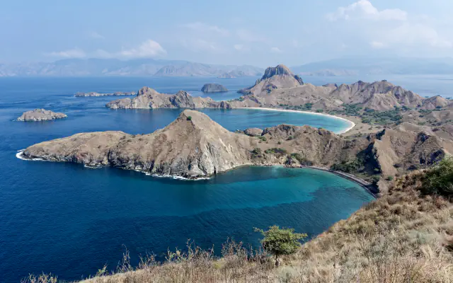 A curved bay of turquoise water below dry brown hills on Padar Island, with small rocky islands and a pale beach in the distance, seen from a viewpoint above