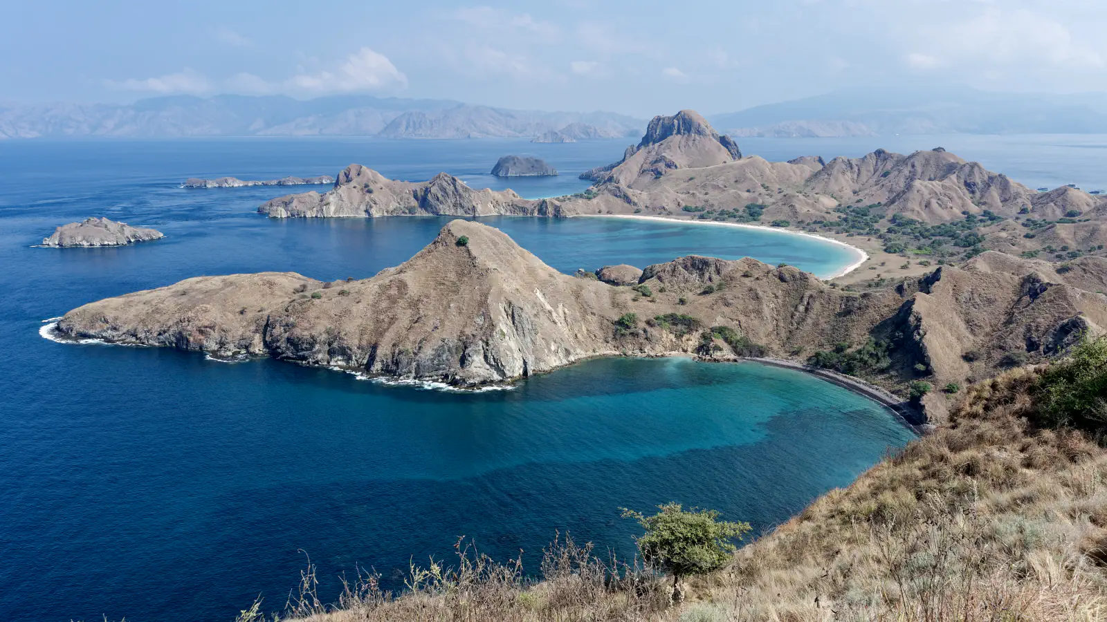 Zavit zaliv turkizne vode pod suhimi rjavimi griči na otoku Padar; v ozadju so manjši skalnati otočki in svetla plaža.