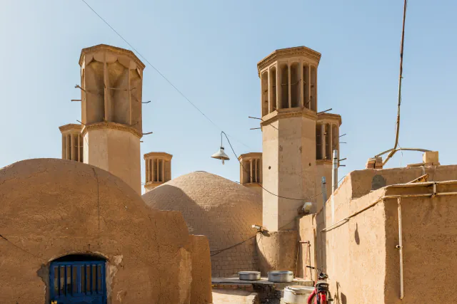 Two prominent wind towers rise above earthen walls and domed roofs in Yazd; several smaller towers appear behind them under a clear sky.
