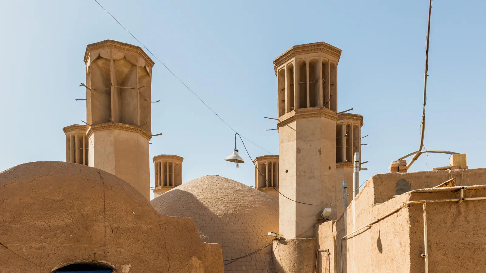 Two prominent wind towers rise above earthen walls and domed roofs in Yazd; several smaller towers appear behind them under a clear sky.