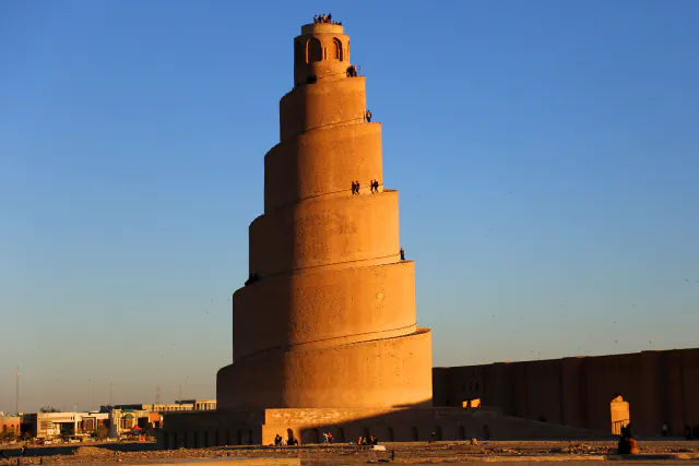 A tall brick spiral minaret with a ramp winding up its tapering tiers to a small open lantern, lit orange by low sun, beside a long brick wall under a clear blue sky