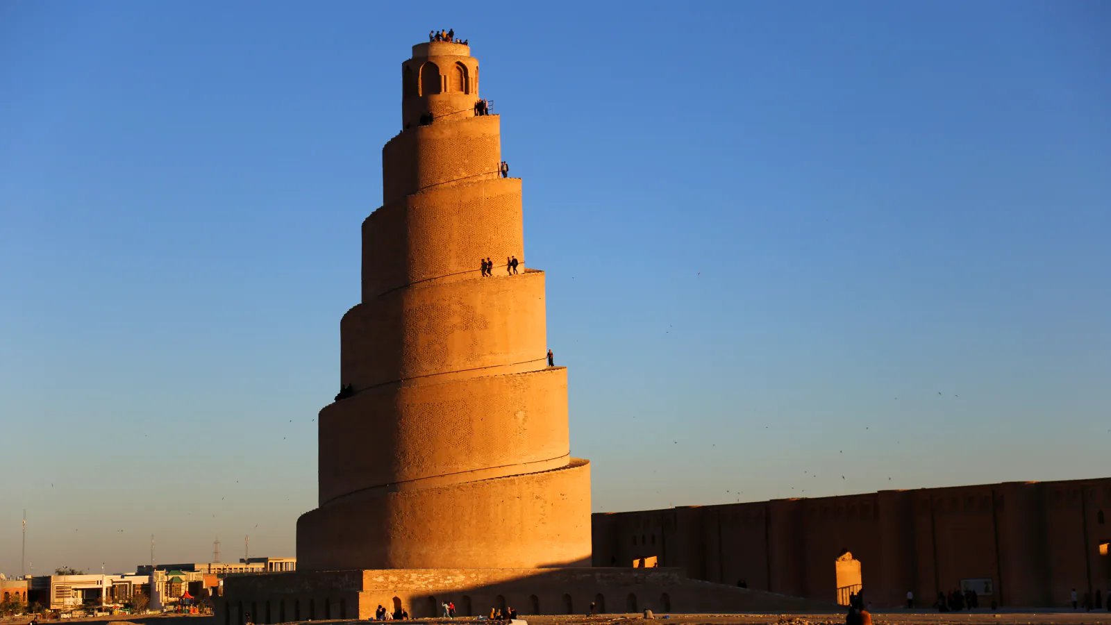 A tall brick spiral minaret with a ramp winding up its tapering tiers to a small open lantern, lit orange by low sun, beside a long brick wall under a clear blue sky