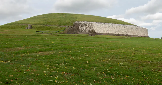 S travo porasla gomila Newgrange s svetlim kamnitim zidom ob vznožju, vidna čez trato pod oblačnim nebom.