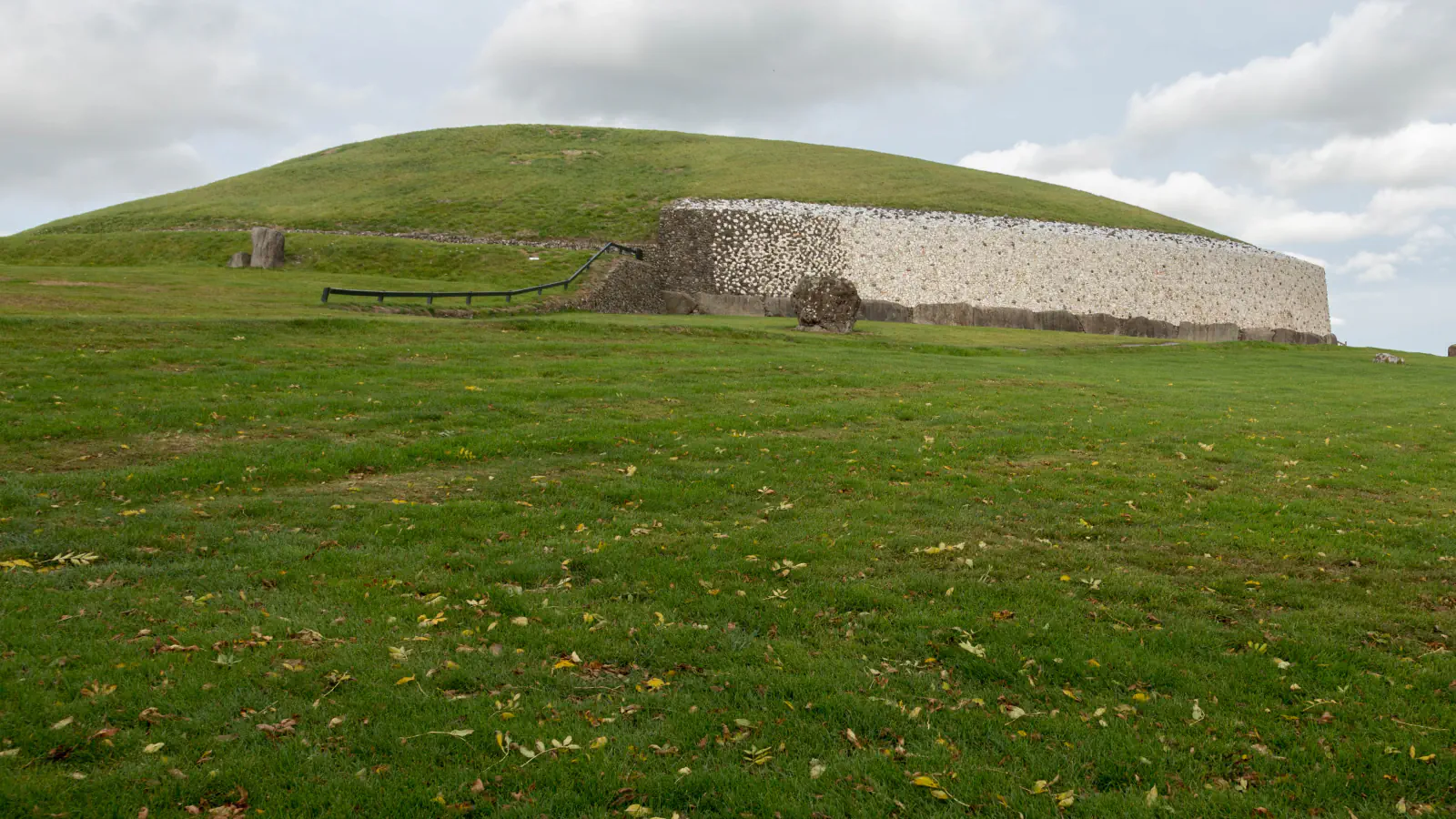 Grass-covered Newgrange mound with a pale stone wall along its base, seen across a lawn under cloud