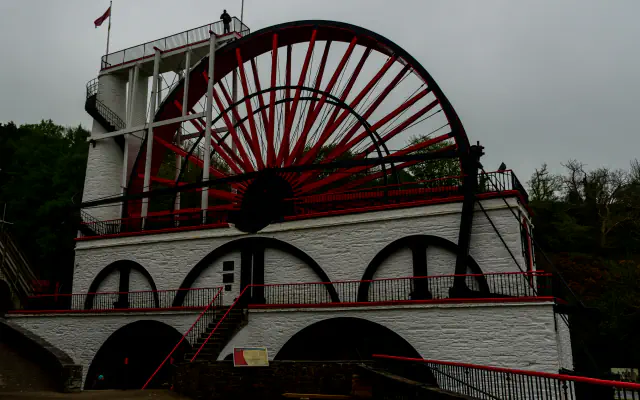 The Great Laxey Wheel on the Isle of Man: a large wheel with red spokes above white-rendered stone arches, a viewing platform on the left, dark trees behind and an overcast sky