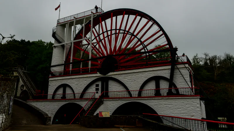 The Great Laxey Wheel on the Isle of Man: a large wheel with red spokes above white-rendered stone arches, a viewing platform on the left, dark trees behind and an overcast sky