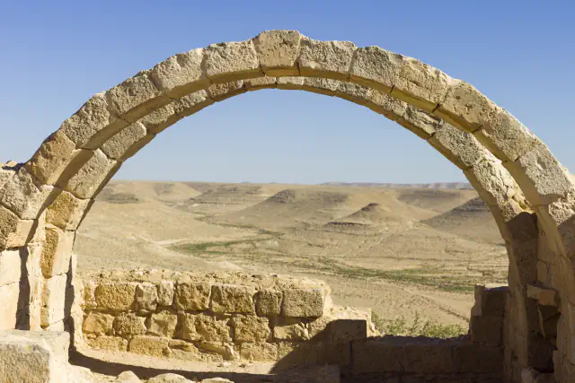 A weathered arch of pale cut stone frames a view of dry, sand-coloured hills under a clear blue sky, with low walls of dressed stone below the arch