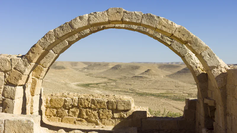 A weathered arch of pale cut stone frames a view of dry, sand-coloured hills under a clear blue sky, with low walls of dressed stone below the arch
