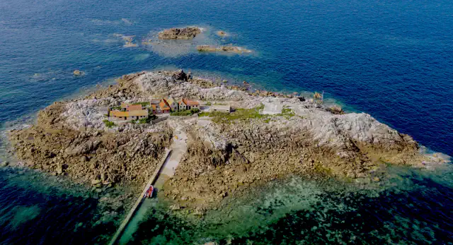Pogled iz zraka na La Maîtresse Île v Les Minquiers: skalnat otoček z vrsto majhnih hiš in pomolom, obdan z vodo in temnejšimi grebeni