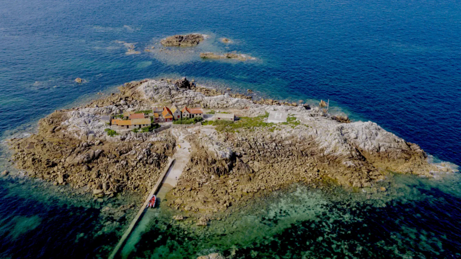 Pogled iz zraka na La Maîtresse Île v Les Minquiers: skalnat otoček z vrsto majhnih hiš in pomolom, obdan z vodo in temnejšimi grebeni