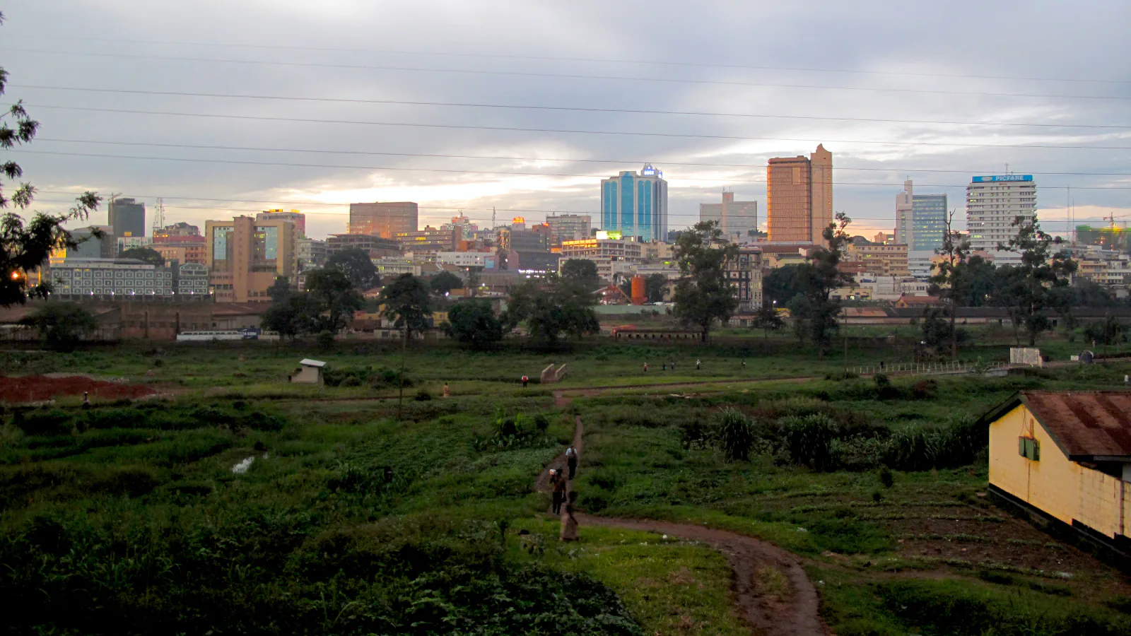 Kampala’s towers and buildings beyond green open ground and a footpath under a cloudy sky.