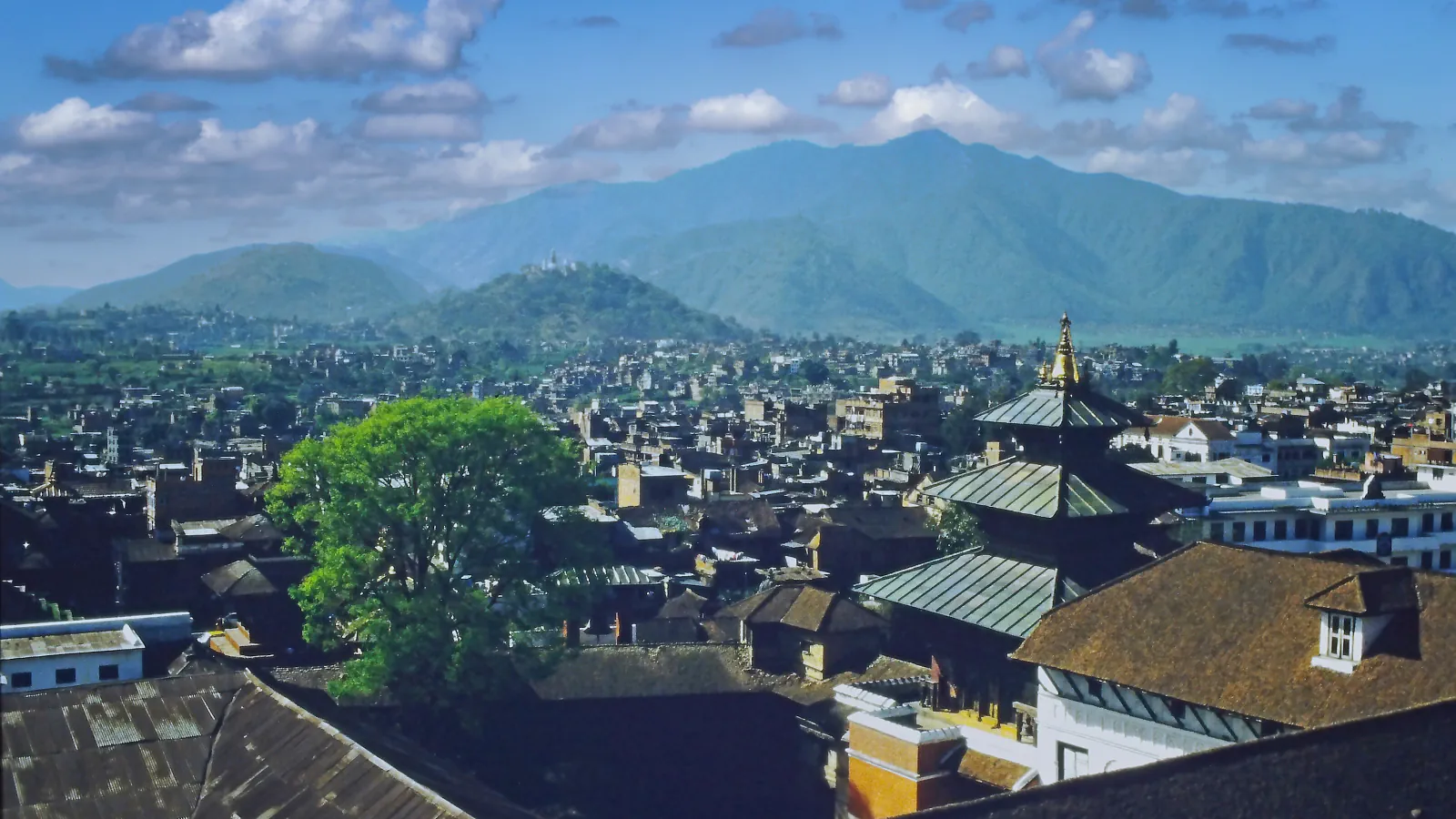 Kathmandu rooftops below wooded hills under a partly cloudy sky; view dated 1987