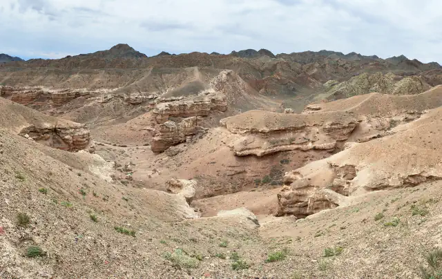 Wide view into a dry valley of layered pink and beige slopes, with gullies and rock ledges below dark ridges under a cloudy sky