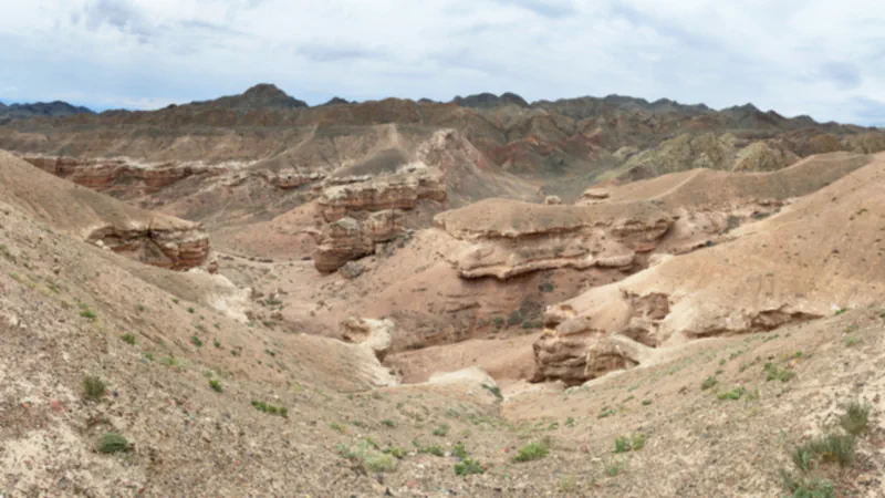 Wide view into a dry valley of layered pink and beige slopes, with gullies and rock ledges below dark ridges under a cloudy sky