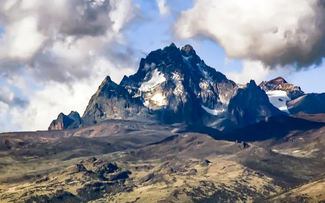 Dark, jagged rock summits of Mount Kenya with patches of snow and ice among large clouds, above grassy and rocky slopes