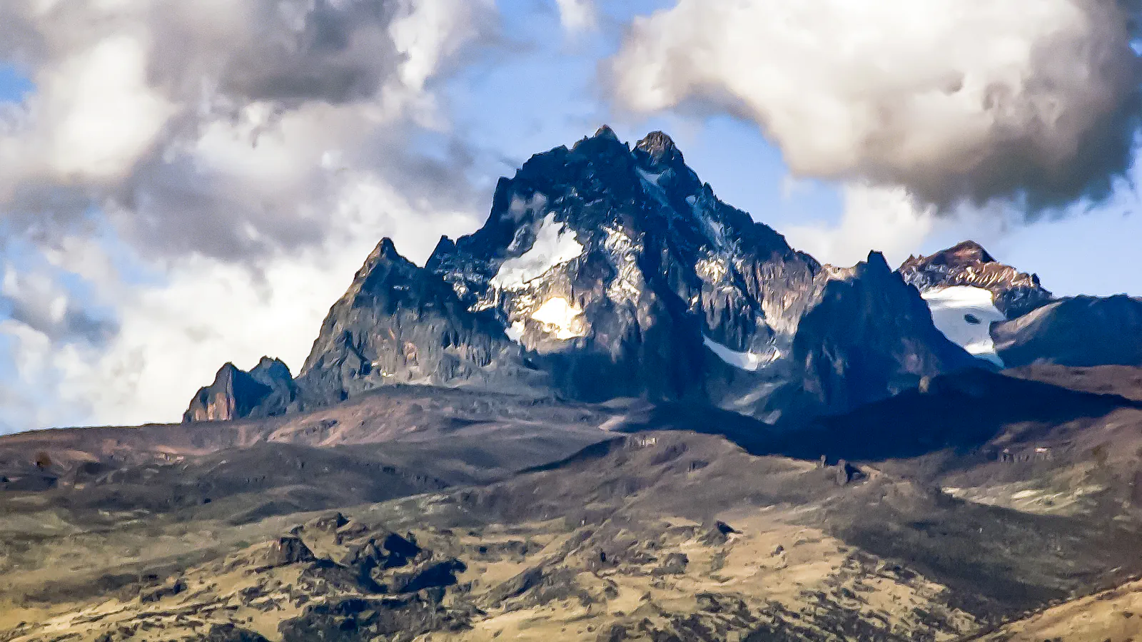 Dark, jagged rock summits of Mount Kenya with patches of snow and ice among large clouds, above grassy and rocky slopes