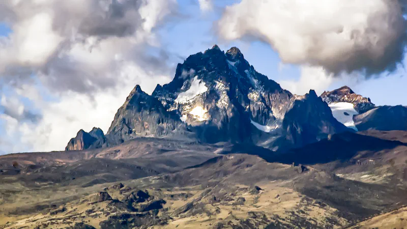 Dark, jagged rock summits of Mount Kenya with patches of snow and ice among large clouds, above grassy and rocky slopes