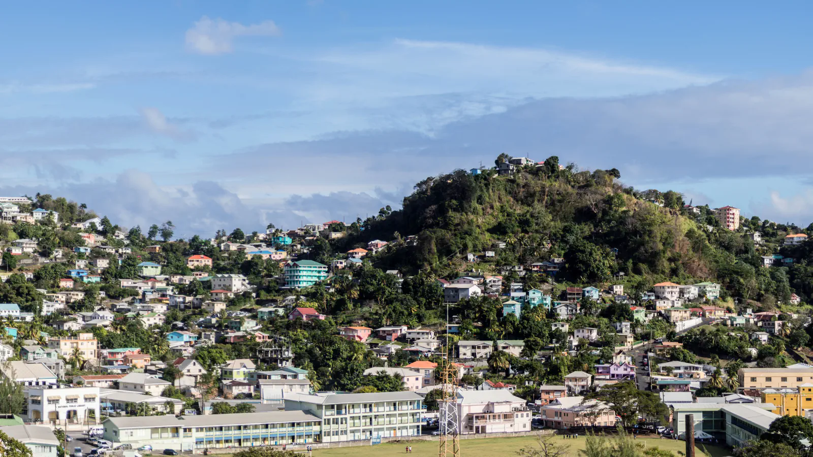 Houses climbing a green hill above central Kingstown beneath a blue, cloudy sky