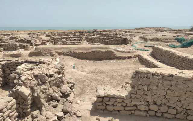 Low walls of stacked stone form roofless rooms across a sandy excavation site, with a strip of blue-green sea on the horizon under a pale sky