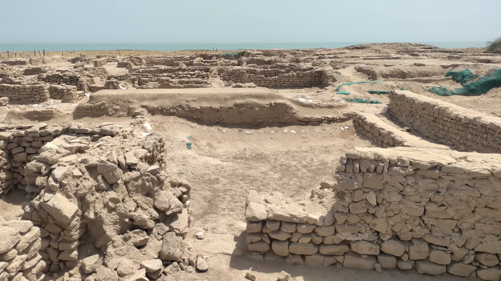 Low walls of stacked stone form roofless rooms across a sandy excavation site, with a strip of blue-green sea on the horizon under a pale sky