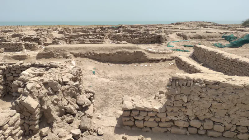 Low walls of stacked stone form roofless rooms across a sandy excavation site, with a strip of blue-green sea on the horizon under a pale sky