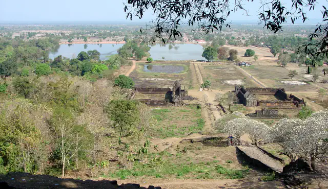 View east from the slope of a mountain at Vat Phou: a straight causeway runs between the ruined walls of two rectangular stone buildings towards a pond and two large reservoirs, with farmland beyond and trees in white blossom at the right