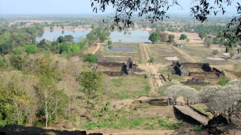 View east from the slope of a mountain at Vat Phou: a straight causeway runs between the ruined walls of two rectangular stone buildings towards a pond and two large reservoirs, with farmland beyond and trees in white blossom at the right