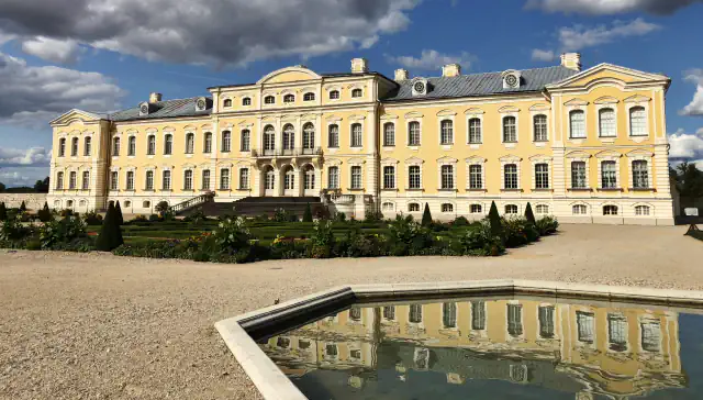 Yellow and white façade of Rundāle Palace seen across a gravel court and a clipped garden, with a rectangular pool in the foreground reflecting the building under dark clouds and blue sky