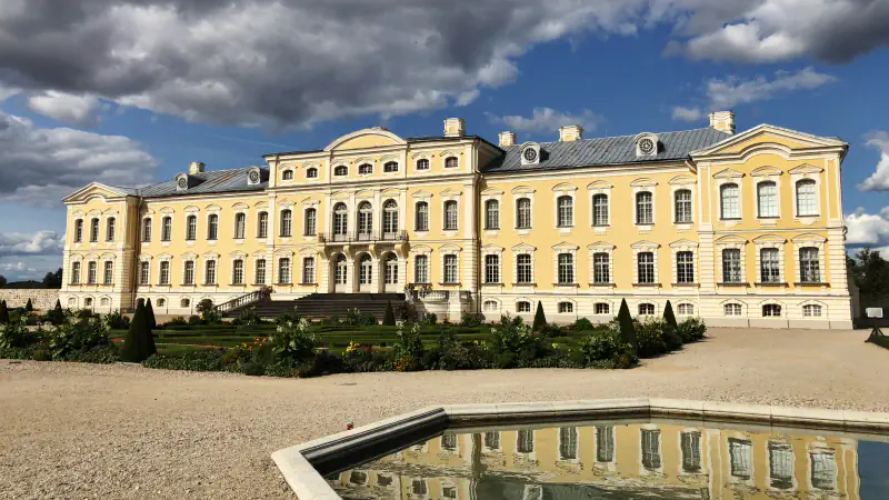 Yellow and white façade of Rundāle Palace seen across a gravel court and a clipped garden, with a rectangular pool in the foreground reflecting the building under dark clouds and blue sky