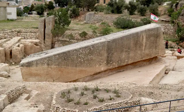 A huge rough-cut stone monolith in the Baalbek quarry, with a Lebanese flag on top, two people beside it, and low stone walls and plants around the site.