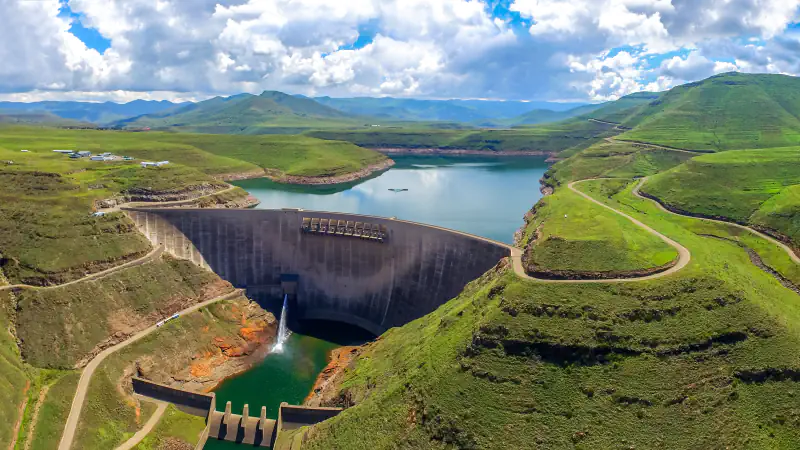 A curved concrete arch dam closes a narrow green valley with a reservoir behind it, a thin jet of water falls from the dam into a green pool below, and grassy hills rise under white cloud