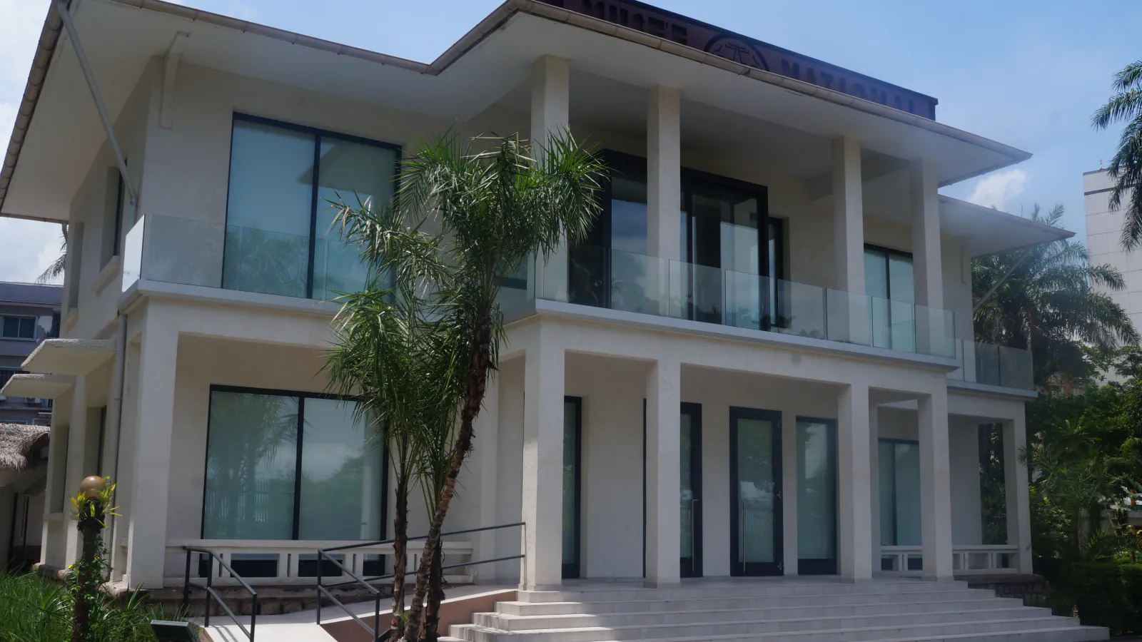 White museum building with two visible storeys, an open upper balcony with a glass railing, broad steps, palms and a museum sign along the roof edge.