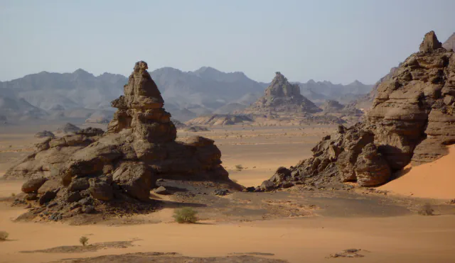 Weathered brown rock towers on a desert plain: a tall rock tower on dark boulders at the left, a rocky ridge with an orange dune at the right, flat sand between them and grey mountain ranges in the haze behind.