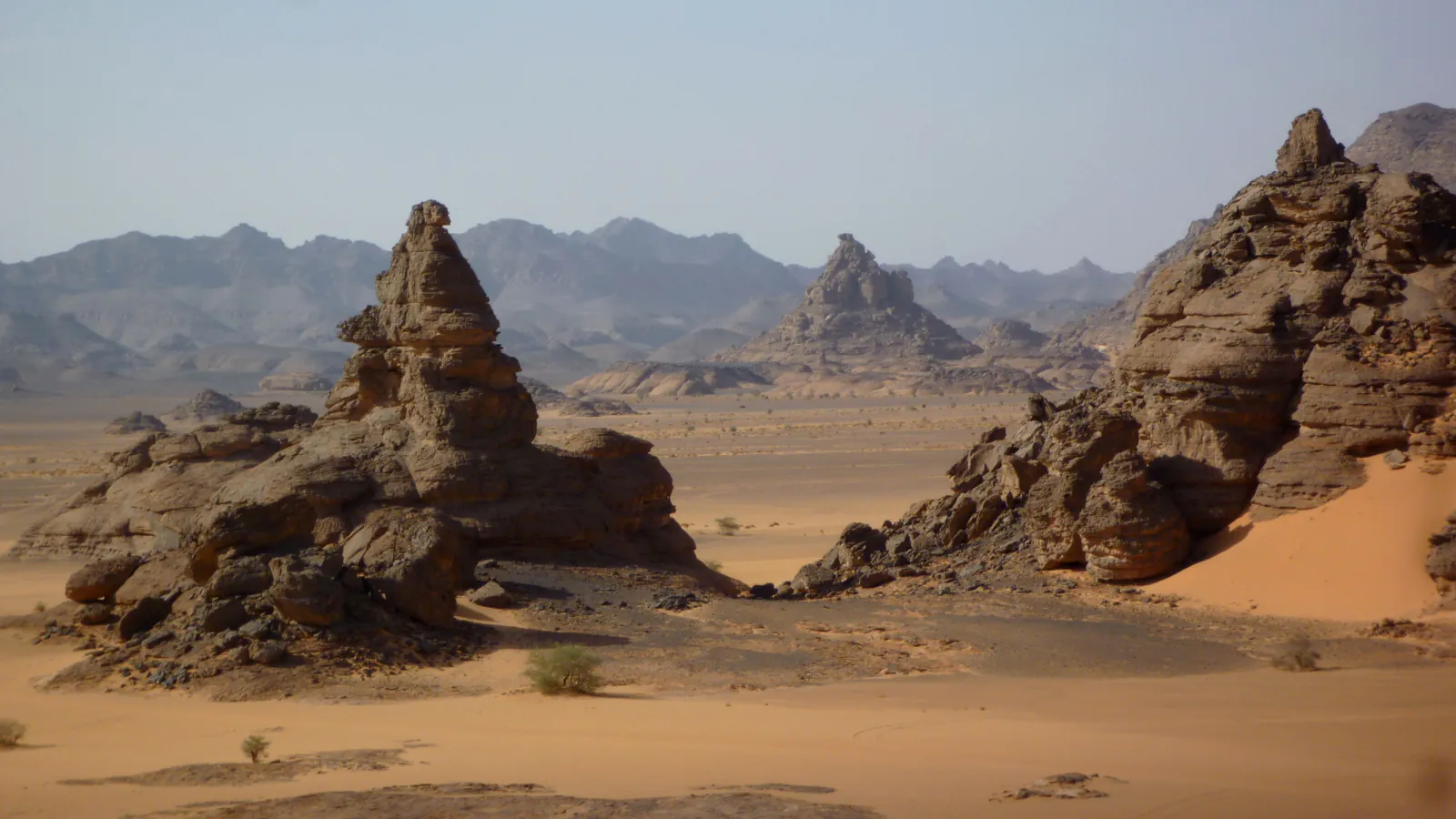 Weathered brown rock towers on a desert plain: a tall rock tower on dark boulders at the left, a rocky ridge with an orange dune at the right, flat sand between them and grey mountain ranges in the haze behind.
